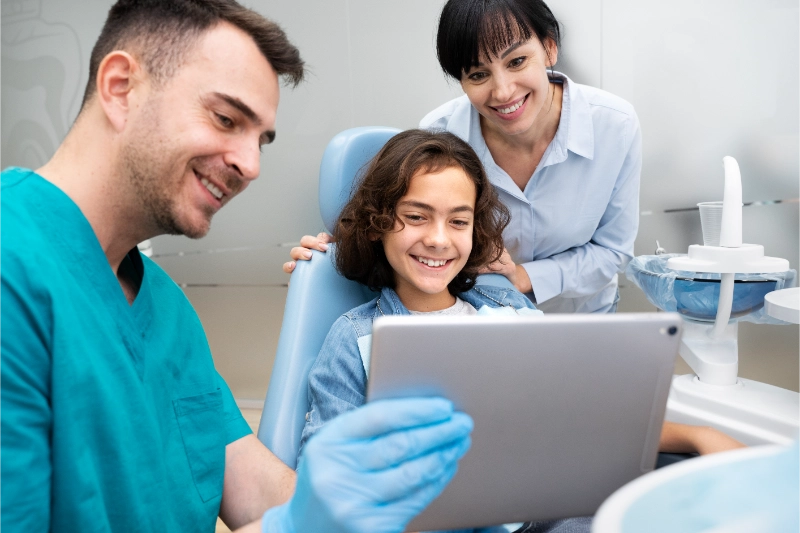 Dentist showing a tablet to a smiling child patient, with a supportive parent nearby, in a dental office setting.