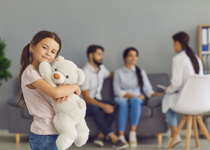 Smiling girl holding a stuffed teddy bear in a pediatric dental office, with parents and a dentist in the background, emphasizing a friendly and comforting environment for children's dental visits.
