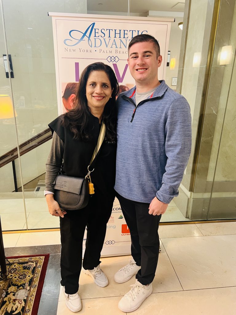 Woman in black attire and man in blue sweater standing together in front of an Aesthetic Advantage banner, emphasizing cosmetic dentistry and smile enhancement.